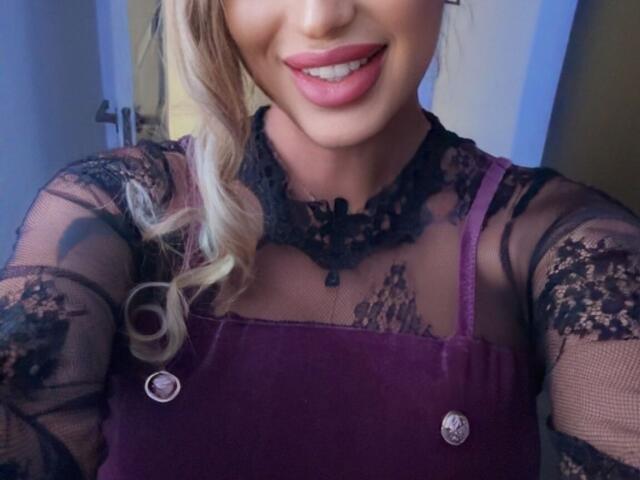 NicolSNaya with blonde hair, pink lips, wearing a sheer black lace top and purple velvet apron, showing a cute smile and her tits through the lace.