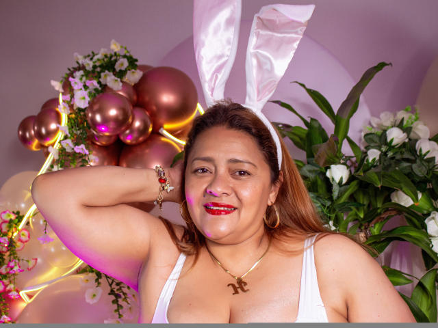 MonzerratBoyer with brown hair, white bunny ears, red lipstick, hoop earrings, necklace, white tank top, smiling, arm raised, pink and gold balloons, flowers, and green plants in background.