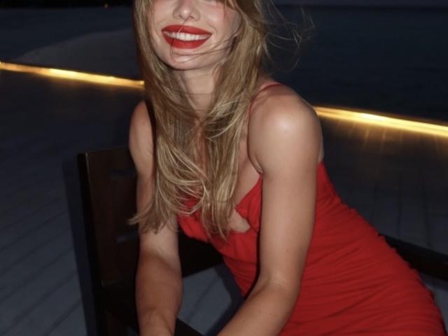TheHottestBlonde in a red dress, leaning forward, showing her tits, with long blonde hair, red lipstick, and a sexy smile, in a dimly lit room.
