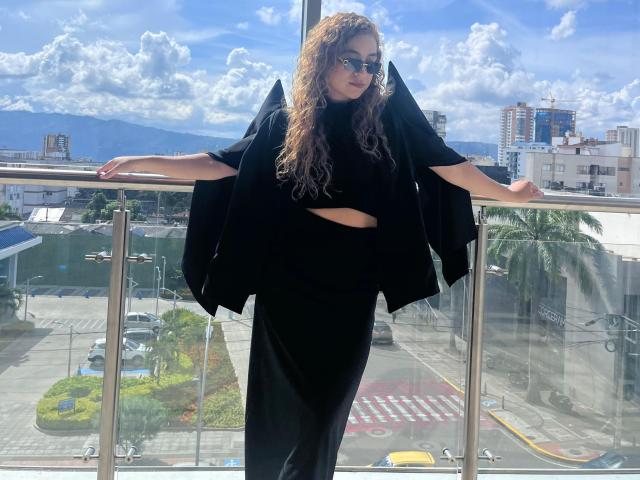 AriannaBlake with curly brown hair, wearing black sunglasses, black crop top, and black pants, standing on a balcony, arms out, black cape draped over shoulders, sunny day.