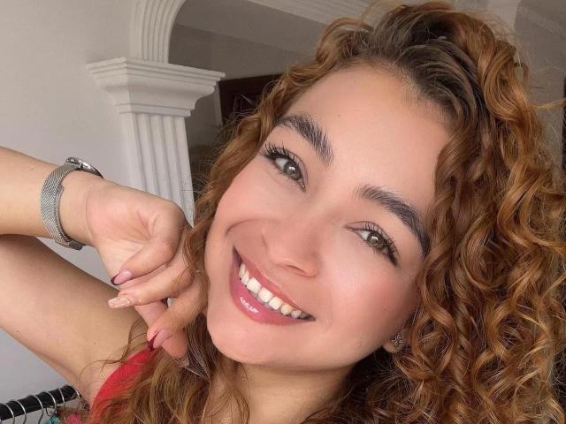 Close-up photo of AriannaBlake with curly brown hair, smiling at the camera. She has light skin, green eyes, and is wearing a red sleeveless top. Her right arm is raised, showing a silver wristwatch and manicured nails with pink polish. Her eyebrows are thick, and she has long eyelashes. The background includes a white archway. The composition focuses on her face and upper torso. She has a shiny lip gloss and a relaxed, happy expression.