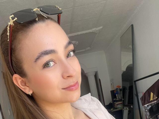 Arianna Blake, con i capelli castani raccolti in una coda di cavallo, occhi verdi, un leggero rossetto rosa e un sorriso delicato, indossa occhiali da sole sulla testa, una camicia bianca e piccoli orecchini..