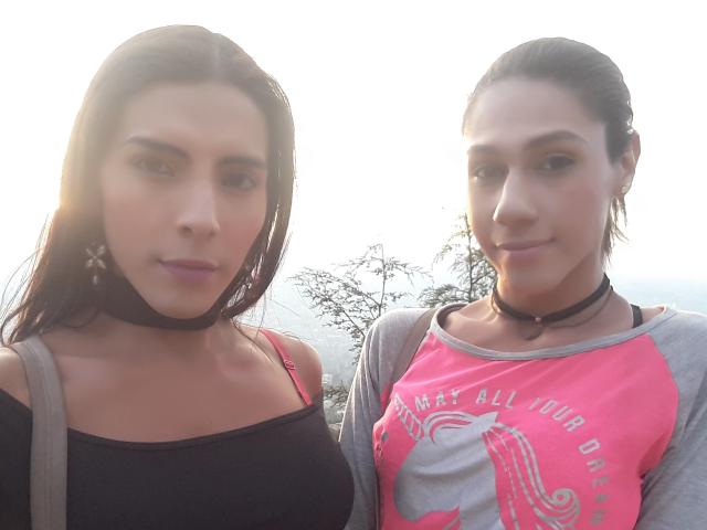 Two women with dark hair, both wearing chokers. AndreaTatyTs on the left has a black strap top, red strap visible, and a cross earring. AndreaTatyTs on the right wears a pink and gray raglan shirt with a white elephant graphic. Both have light skin and subtle makeup. Soft sunlight from the back. The woman on the right has a slight smile, while the woman on the left looks neutral. The image is a selfie, slightly overexposed.