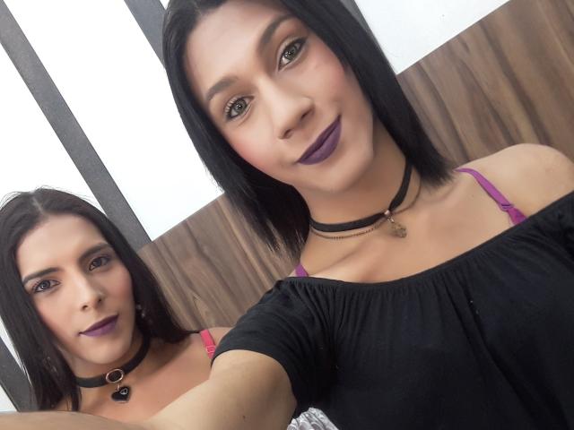Two portraits of AndreaTatyTs with straight black hair and purple lipstick. She wears a black off-shoulder top with pink straps, a black choker with a small heart, and a gold pendant. Her skin is light, and she has dark, defined eyebrows. The foreground portrait is closer, showing her face and upper body, while the background portrait is slightly smaller and shows her from a three-quarter angle. Both portraits have a neutral, indoor setting.