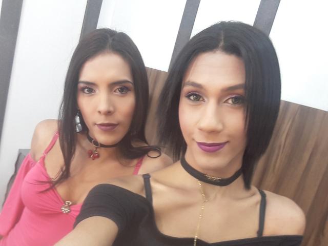 AndreaTatyTs with dark hair, purple lipstick, black choker, pink top, showing boobs, and black top, holding phone, standing next to AndreaTatyTs with same makeup and choker.