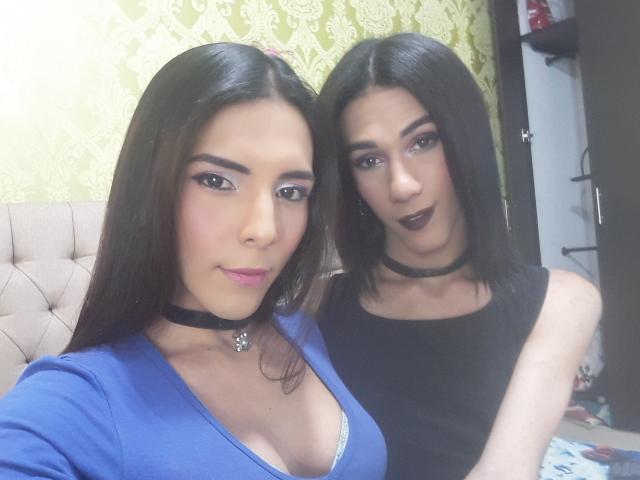 AndreaTatyTs with long black hair, pink lipstick, blue top showing big tits, black choker, next to AndreaTatyTs in black top, dark lipstick, black choker, in bedroom.