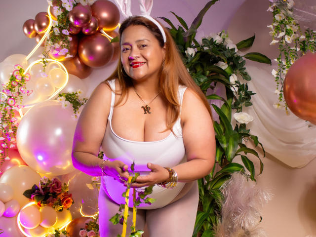 MonzerratBoyer with medium brown skin, large boobs, white tank top, bunny ears, smiling, holding green ribbon, surrounded by pink and gold balloons, white flowers, and green plants.