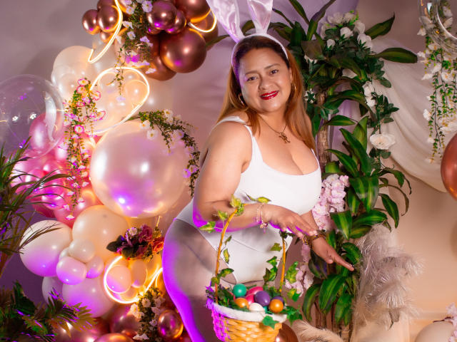 MonzerratBoyer with bunny ears, white tank top, and white panties, holding a basket of colorful eggs, smiling, red lipstick, medium tits, standing among balloons and plants.