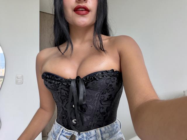 EmilyJensenn with dark black hair, red lipstick, wearing a black lace corset, showing her large, round tits, and denim jeans, taking a selfie in a well-lit room.