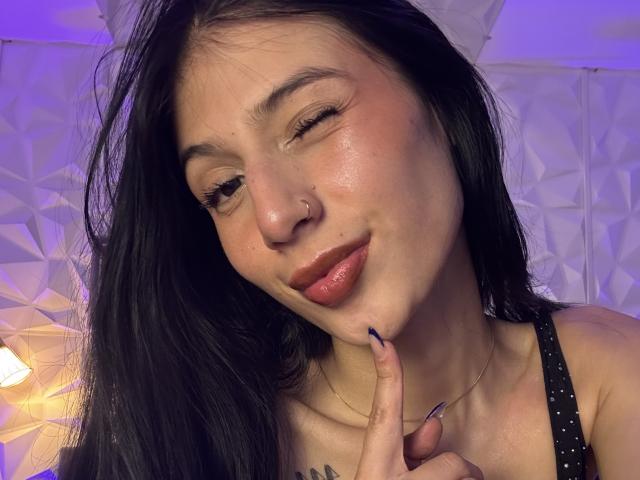 SophieBeaumont with black hair, nose ring, and pink lipstick, winking and touching her chin with a finger, wearing a black sleeveless top, showing a cute smile.