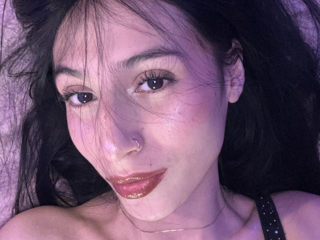 Close-up of SophieBeaumont's face with dark, slightly wavy hair covering her forehead and eyes. She has fair skin, subtle makeup with dark red lipstick, and a small nose ring on her left nostril. Her eyes are dark brown, and she wears a thin silver necklace. She has a confident, slightly smirking expression. The image has a purple-blue tint, adding a cool tone to the overall composition. Her black hair contrasts with her light skin and dark lipstick. The texture of her hair is soft and slightly messy.