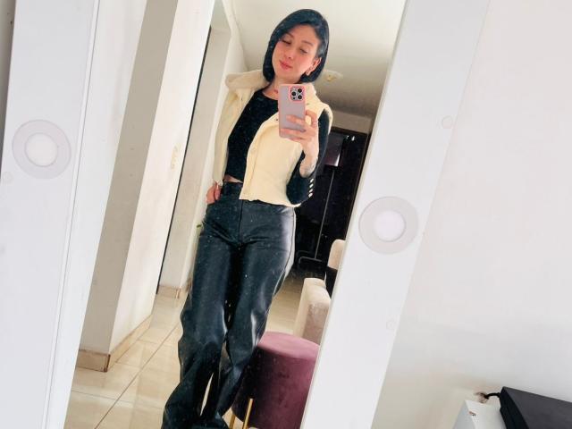 Valeriaaf stands in front of a mirror, wearing a black hoodie, yellow vest, and dark jeans, holding a pink phone, smiling, with a cute ass and small tits.