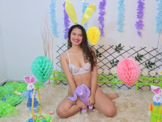 SanetPerlas, con cabello castaño rizado, sonriendo, vestida con un sujetador blanco de encaje, sentada en una alfombra esponjosa, sosteniendo un conejo de peluche morado, rodeada de decoraciones de Pascua, incluyendo farolillos de papel de colores..