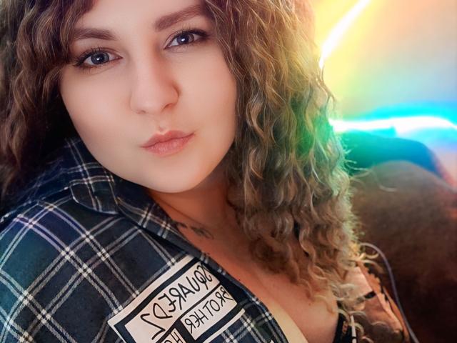 MinistraX with curly brown hair, light skin, and blue eyes, wearing a plaid shirt, has a nose ring and subtle makeup, looking directly at the camera with a pout.