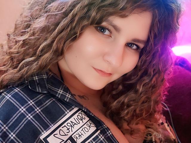 MinistraX with curly brown hair, blue eyes, and a slight smile, wearing a plaid shirt, showing a small tattoo near the collarbone, and a sexy, relaxed expression.