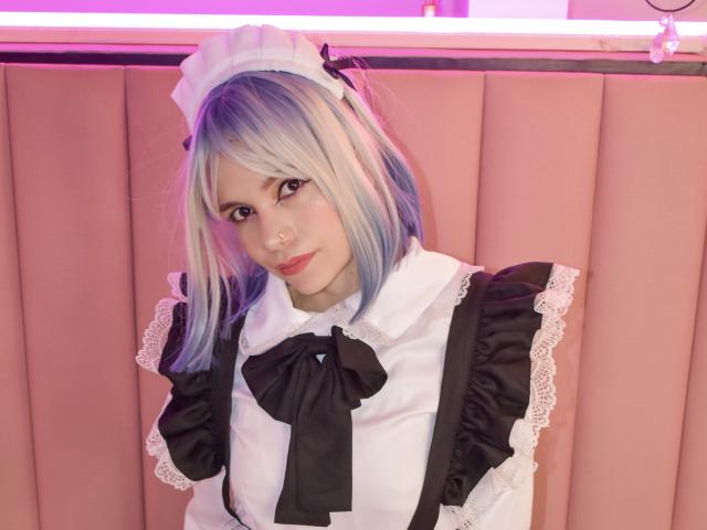 SofiArias with blue and white hair, wearing a black and white maid outfit, lace cuffs, and a black ribbon, standing in front of a pink, padded wall.