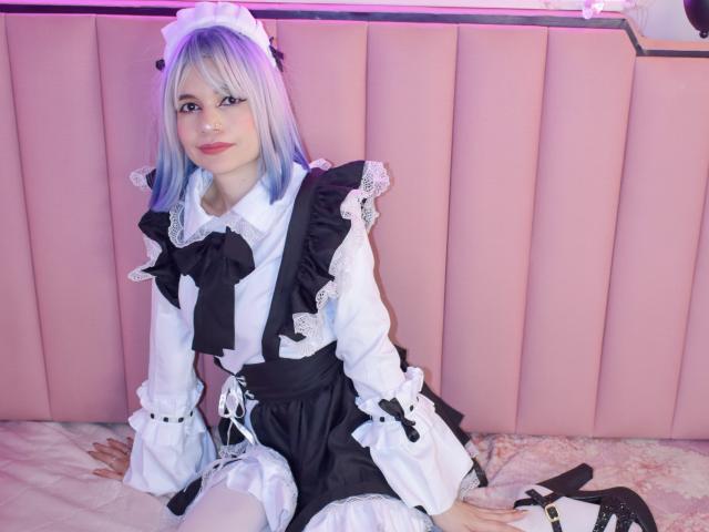 SofiArias with blue and white hair, wearing a black and white maid outfit, sitting on a bed, white thigh-highs, black heels, pink cushioned wall, and a cute smile.