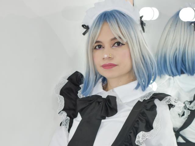 Sofi Arias, con cabello azul y blanco, vestida con un traje de sirvienta negro y blanco con encaje, un lazo negro y un tocado blanco, tiene lápiz labial rosa y un piercing en la nariz..