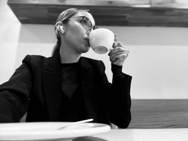 Black-and-white photo of AdharaCooperr sipping from a white mug. AdharaCooperr wears a dark, puffy-sleeve jacket and dark shirt. She has glasses with a decorative pattern and small earrings. Her hair is tied back. The table in the foreground has a white plate with a fork. The wooden shelf in the background is slightly out of focus. The image has a high contrast, emphasizing AdharaCooperr's facial features and the texture of her clothing.