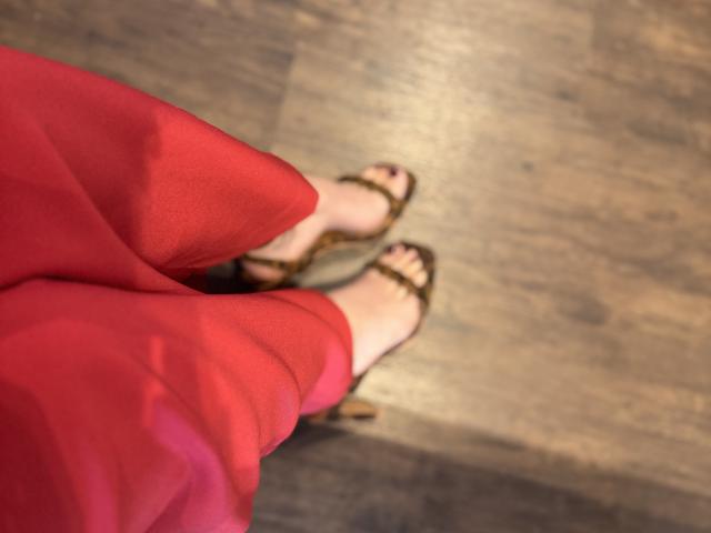 Close-up of AdharaCooperr's feet. AdharaCooperr is wearing a red, semi-sheer dress with their legs slightly apart. AdharaCooperr's feet are in brown strappy sandals, with visible toenails painted purple. The wooden floor has a textured, dark brown pattern. The image focuses on the feet and dress, with a shallow depth of field that blurs the background. The perspective is from above, looking down at AdharaCooperr's feet.