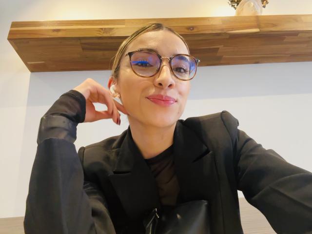 AdharaCooperr has light skin, wears large, round glasses with a slight blue reflection, and has brown hair pulled back. She's dressed in a black, sheer, long-sleeve top with a black blazer. She rests her chin on her right hand, smiling with pink lipstick. A wooden shelf is in the background. Her earrings are small and gold. The image is a selfie.