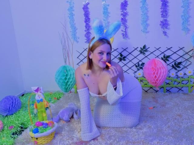 CristalMegan kneels on a fluffy white rug, wearing a white fishnet dress with long sleeves and light blue, furry bunny ears. She has fair skin, red hair, and small tattoos on her left shoulder. She's eating a colorful lollipop, her right arm on the rug. The background features purple and blue paper chains, a green lattice, and a basket with Easter eggs and a plush bunny. The scene is brightly lit with a blue-purple tint.