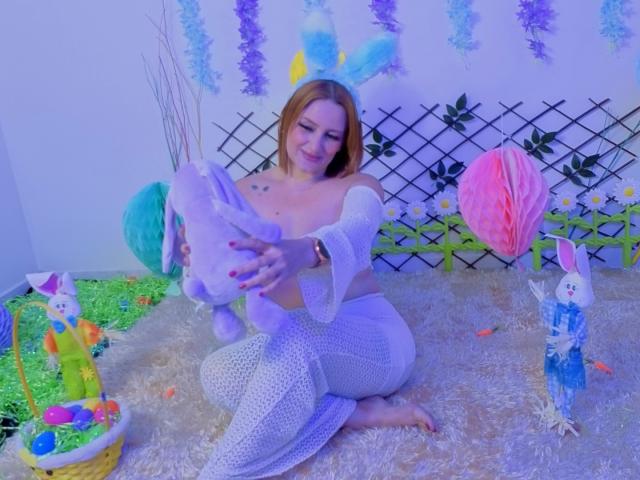 CristalMegan with medium tits, fair skin, and blonde hair, kneeling on a fluffy rug, holding a purple stuffed bunny, wearing white mesh pants and a blue headband with bunny ears.