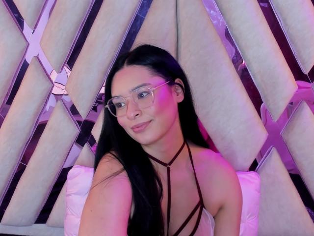 IsaDoux with black hair, glasses, and a strappy top, showing her tits in a selfie, smiling with pink and purple lighting.