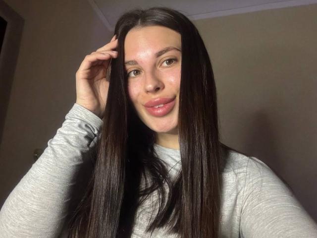 BlackFatal mit langem, dunkelbraunen Haar, einem grauen Hemd, leicht geschminkten, rosafarbenen Lippen und einer Hand im Haar, lächelnd. Helle Haut, braune Augen, dezentes Make-up. Sie ist süß..
