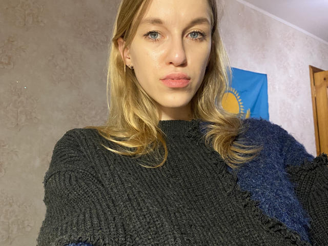 DeedraGalarneau with blonde hair, blue eyes, and pink lips, wearing a black, fuzzy sweater, has her tits slightly visible, looking into the camera with a neutral expression.