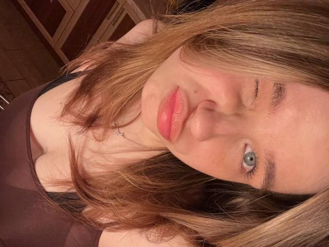 JennyOsmun with winking blue eye, pink lips, and brown hair, wearing a sheer top, showing her tits, in a selfie. Her ass is not visible.