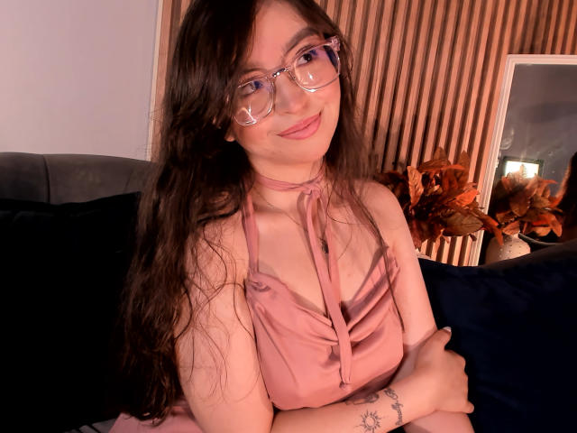 EvaMcAllister with long brown hair, glasses, pink halter dress, small tits, tattoo on right wrist, smiling, arms crossed, sitting on dark blue couch, wooden wall in background.