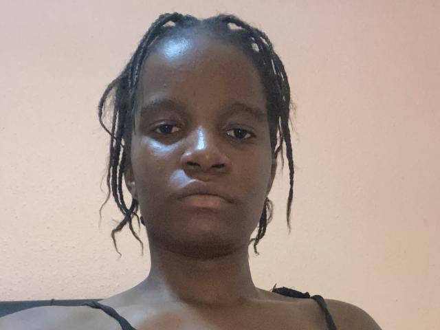MercedesHot has dark skin, small round tits, and is wearing black bra straps. She has short, messy dreadlocks and a serious, neutral expression. Her face is slightly shiny.
