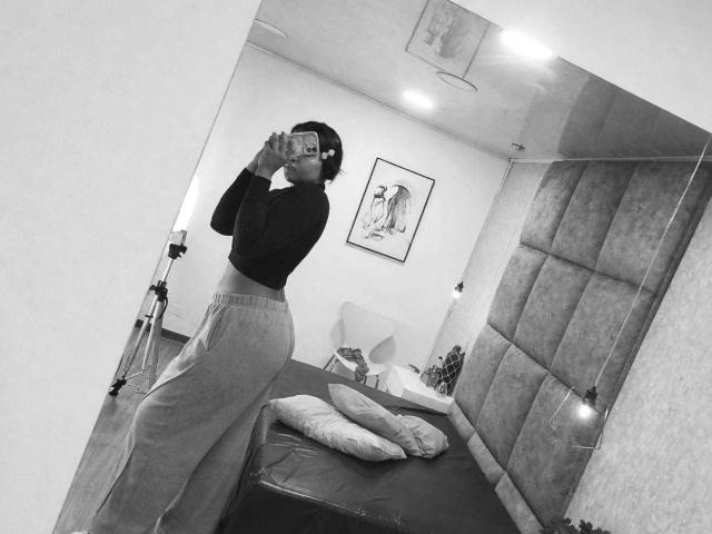 Black-and-white selfie of ChrystalFiore with glasses, wearing a long-sleeve crop top and high-waisted pants, holding a camera, standing by a bed with pillows, and a tripod.