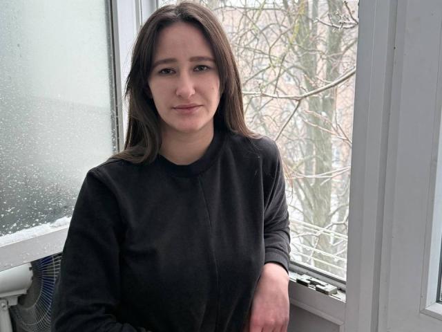 Omella with medium brown hair, wearing a black sweater, has a subtle smile and sits by a window with fogged glass and snow-covered branches outside.