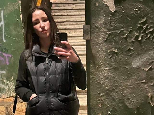 Omella with dark hair, pouty lips, wearing a black puffy jacket, hands in pockets, taking a mirror selfie with a black phone, textured green wall on the right.