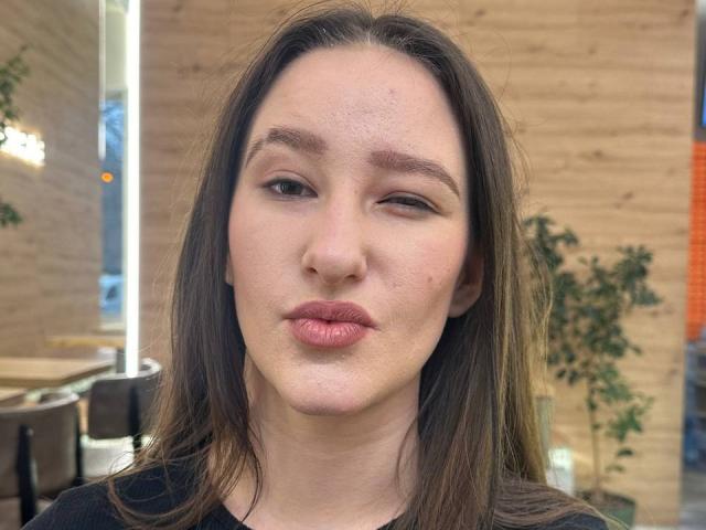 Omella, avec ses lèvres roses et gonflées, cligne de l'œil, a les yeux marron, les cheveux bruns lisses, et quelques petites imperfections sur le front, elle porte une chemise noire, dans une pièce en bois..