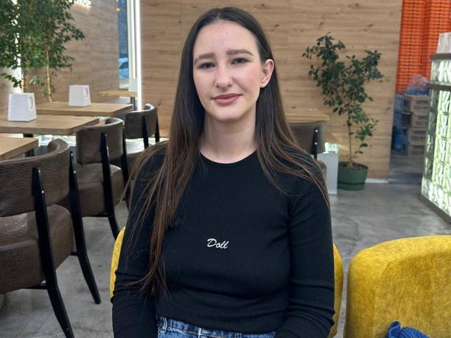 Omella has long brown hair, fair skin, and wears a black long-sleeve shirt with "Dell" on it, sitting in a yellow chair, showing her tits slightly.