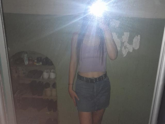 Omella with long brown hair, wearing a purple crop top and blue denim skirt, standing in front of a mirror, flash obscuring face, mirror reflection, belt, bare midriff.