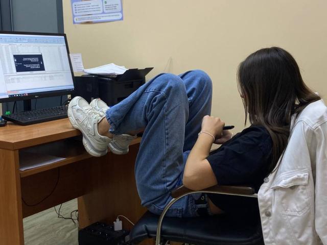 Omella's ass is up in the air, legs crossed on a wooden desk, wearing white sneakers and blue jeans, sitting on a chair in front of a laptop.