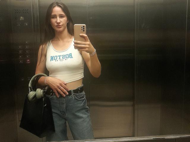 Una mujer con cabello castaño largo, un top blanco que deja ver pequeños senos, jeans azules, un bolso negro y auriculares verdes, tomando un selfie en un ascensor de metal, sosteniendo un teléfono..