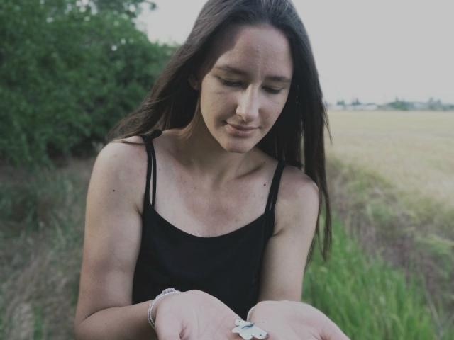 Omella with long brown hair, wearing a black tank top, looking down at something in her hands, smiling slightly, in a grassy field, with a green tree in the background.