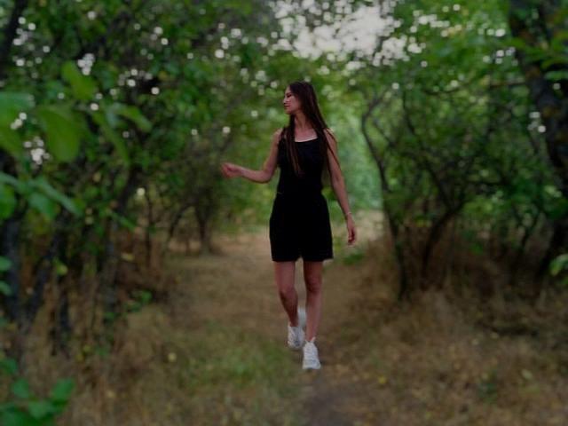 Omella, black dress, white sneakers, running down a dirt path, green trees on both sides, mid-stride, smiling, long brown hair, arms slightly raised, natural light.