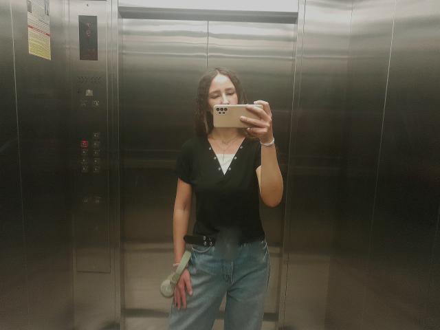 Omella, with medium tits, wearing a black top and blue jeans, takes a mirror selfie in a metal elevator, holding a green cloth in her right hand.