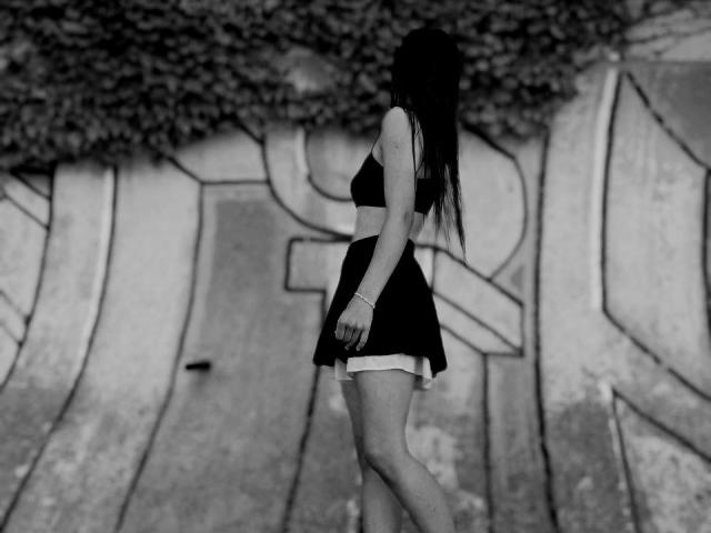 Omella has long dark hair, a black bra, and a short skirt, showing her ass. She's standing sideways, wearing a bracelet, against a graffiti-covered wall.