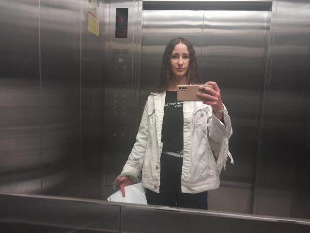 Omella with curly brown hair, white jacket, black shirt, and dark pants, holding a phone and a paper, taking a selfie in a shiny, metal elevator.