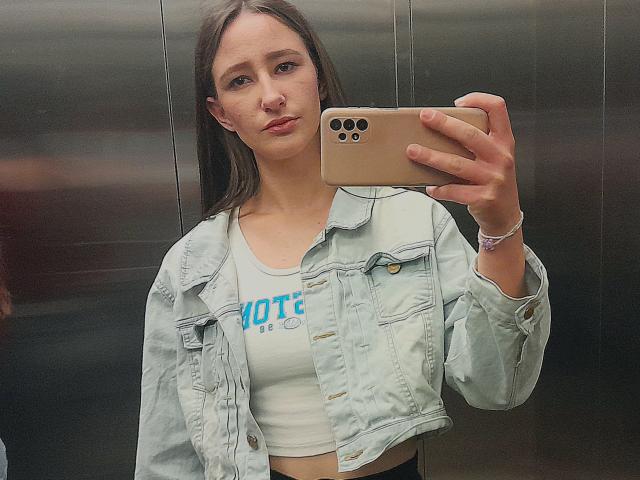 Omella with long brown hair, wearing a light blue denim jacket and white crop top, holding a gold iPhone, taking a mirror selfie with a serious expression.