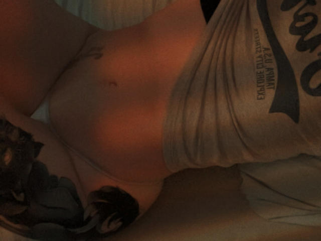 A close-up, dimly lit photograph of AizenMyoo's torso. AizenMyoo is lying on a bed, wearing a grey shirt that's partially pulled up, exposing their lower abdomen and a small tattoo on the right side. AizenMyoo has a visible belly button and is wearing light-colored underwear. A detailed tattoo featuring a dark, intricate design is on the left side of their lower abdomen. The lighting casts warm, reddish tones on AizenMyoo's skin. The shirt has bold, black text on the right.