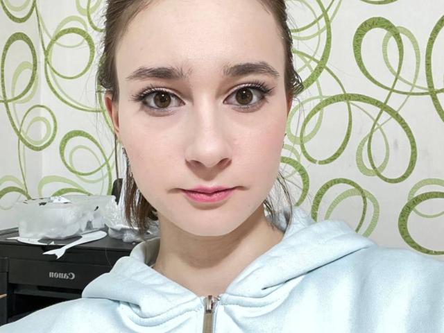 ZofiaWeigold with light skin, brown eyes, and pink lipstick, wearing a light blue hoodie, has small tits and no visible ass, standing in front of a green circular wallpaper.