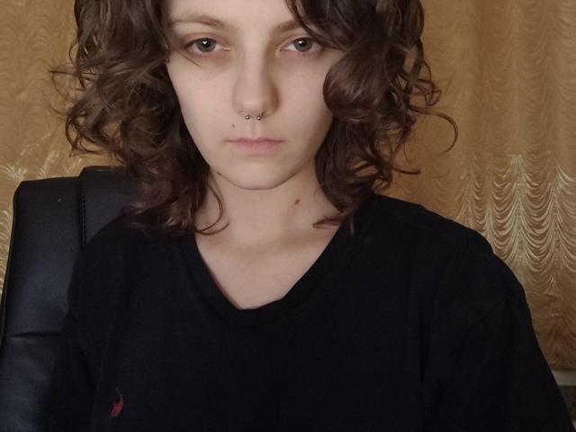 AntionetteRankin with curly brown hair, pale skin, black shirt, nose piercing, and a slight frown, sitting on a black leather chair.